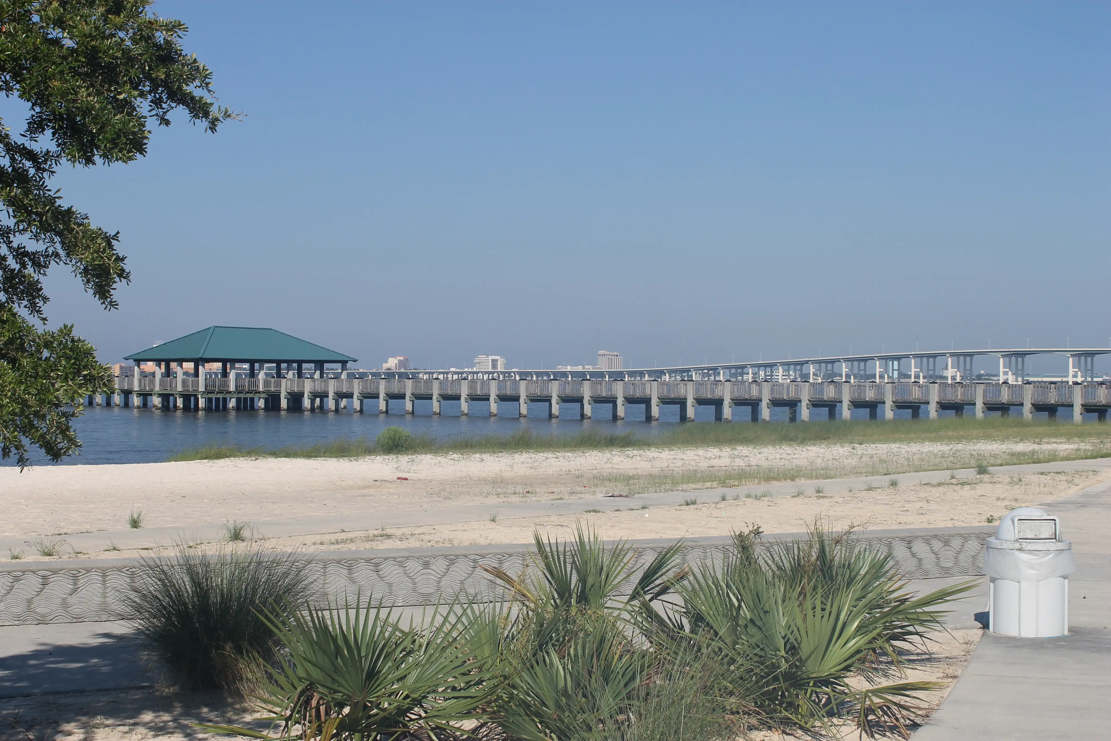 A sandy beach scene with a pier extending out into the water and some low palm shrubs in the foreground