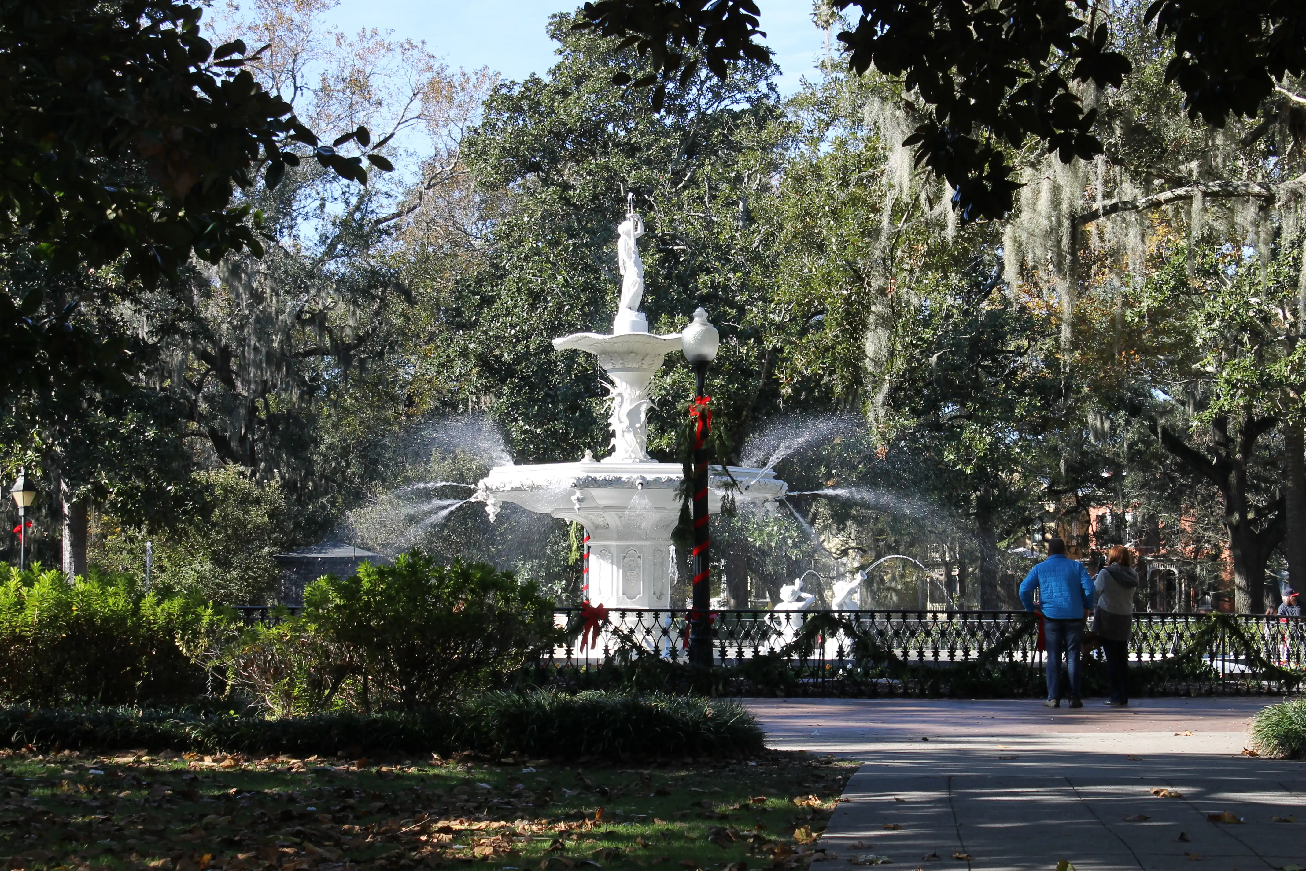 A fountain in a park with a couple taking photos and light posts wrapped in christmas garland