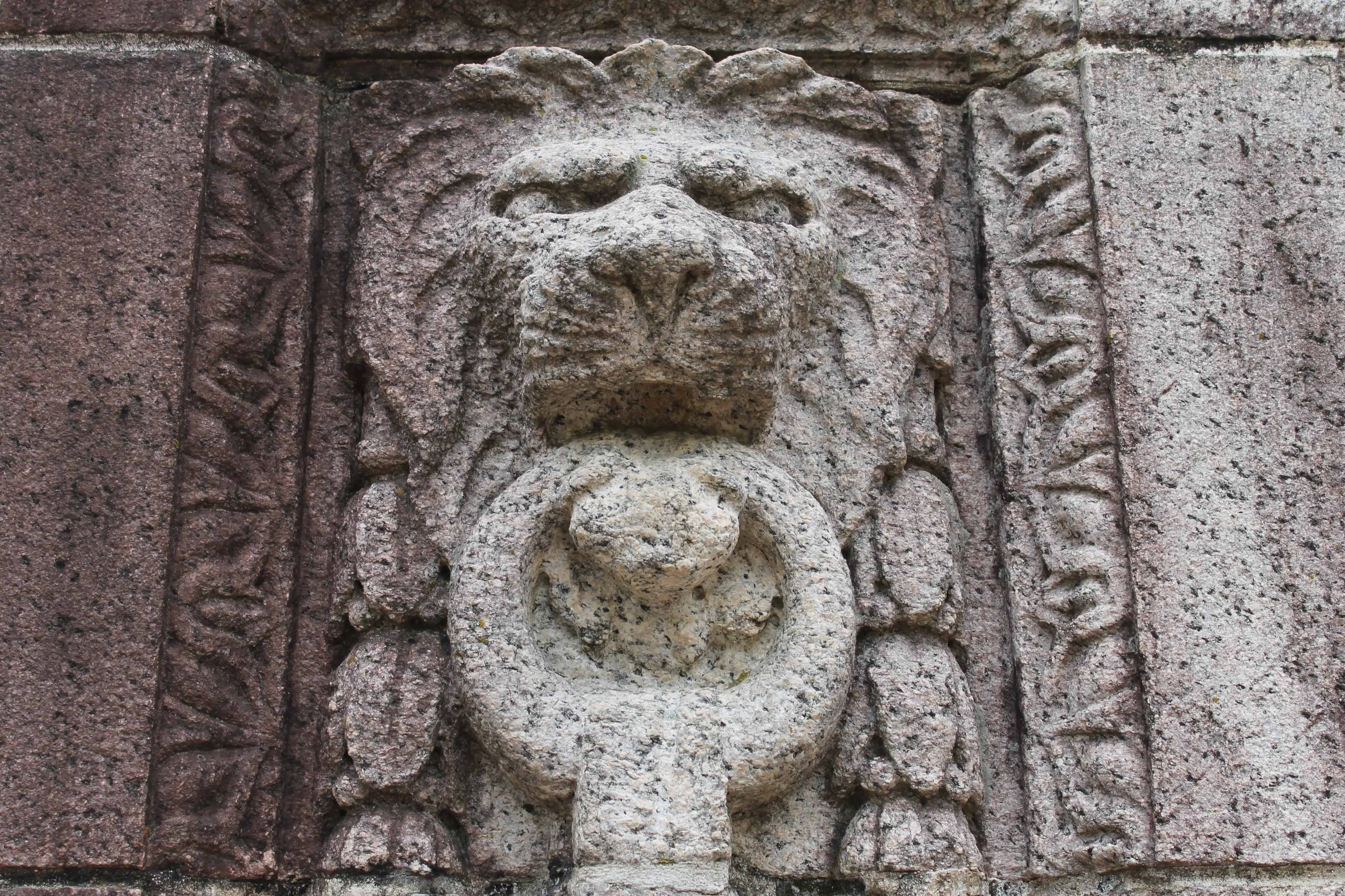 A stone wall carved with a lions head holding a door knocker in its mouth